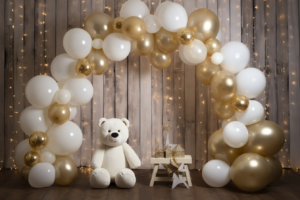 A photo shoot set for a kids' New Year's Eve Smash party. There is a wooden wall in the background and a large, organic arch made of golden and white balloons framing the foreground. The balloons create a whimsical frame. A teddy bear sits below the arch with a "Happy New Year" LED sign hanging above it. Soft natural lighting illuminates the scene. Focal blur creates dreamy bokeh in the background. Extra details and decorations make the scene fun and festive. 8K ultra-high resolution, photorealistic render. --ar 3:2 --stylize 50 --v 5.2